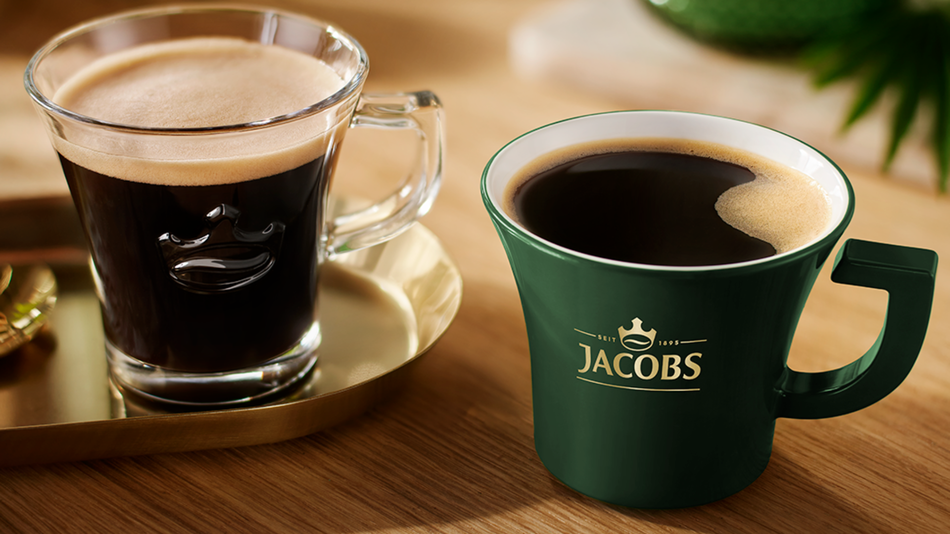 Zwei Kaffeetassen auf Holzoberfläche, eine aus Glas auf goldenem Tablett, eine grüne mit Jacobs-Logo.