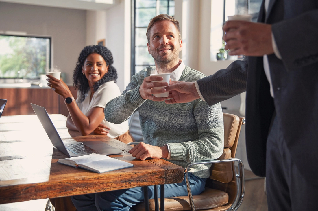Drei Personen in einem modernen Büro, zwei sitzen am Tisch mit Laptops, eine steht und reicht eine Tasse.