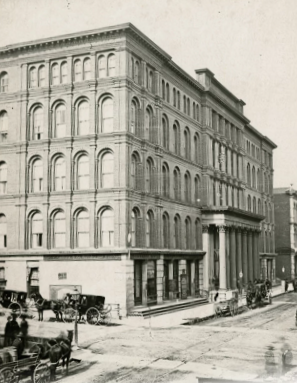 Historisches Gebäude mit Rundbogenfenstern und Pferdekutschen an der Straßenecke.
