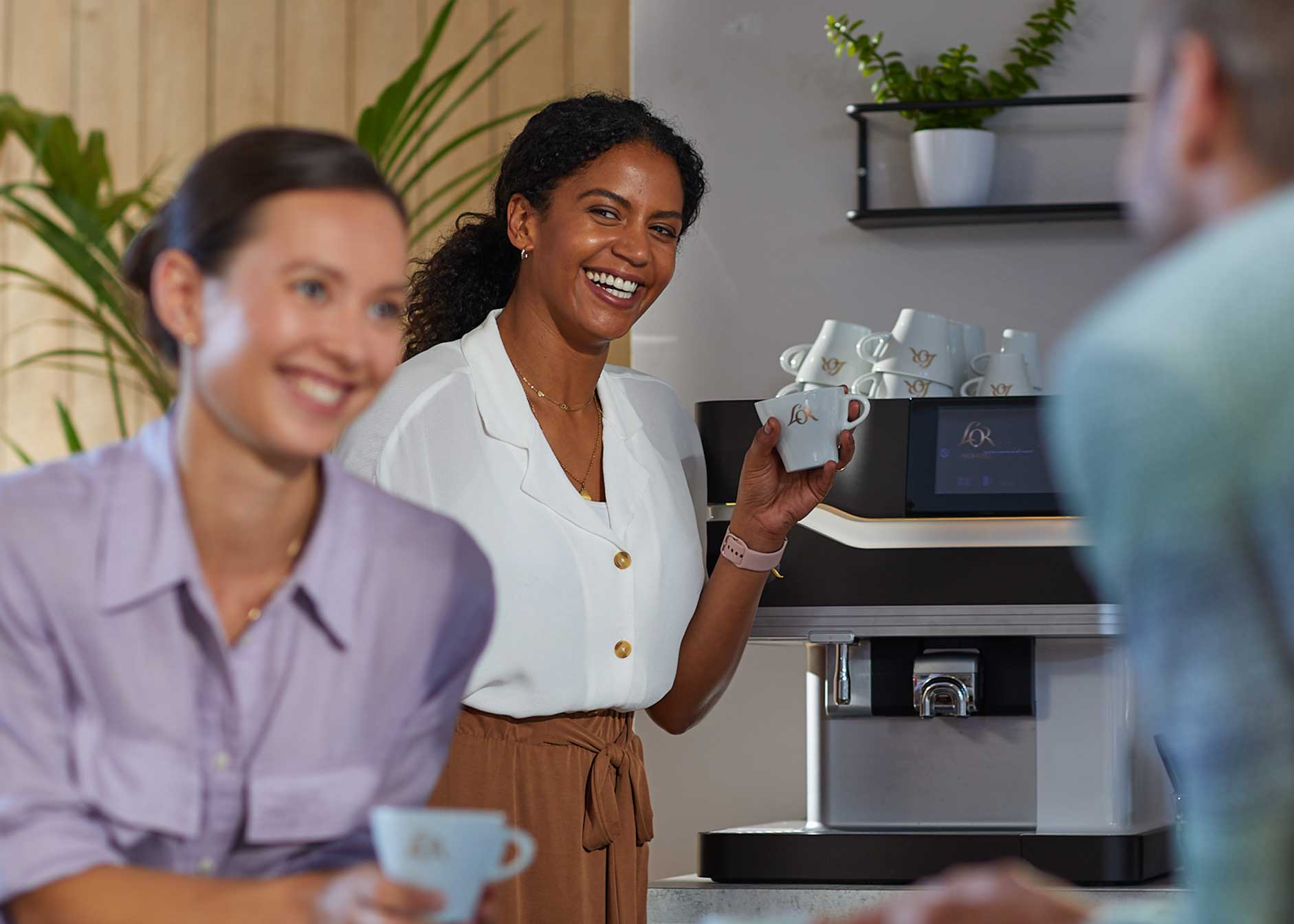 Drei Personen unterhalten sich an einer Kaffeemaschine, jeder hält eine Tasse in der Hand.