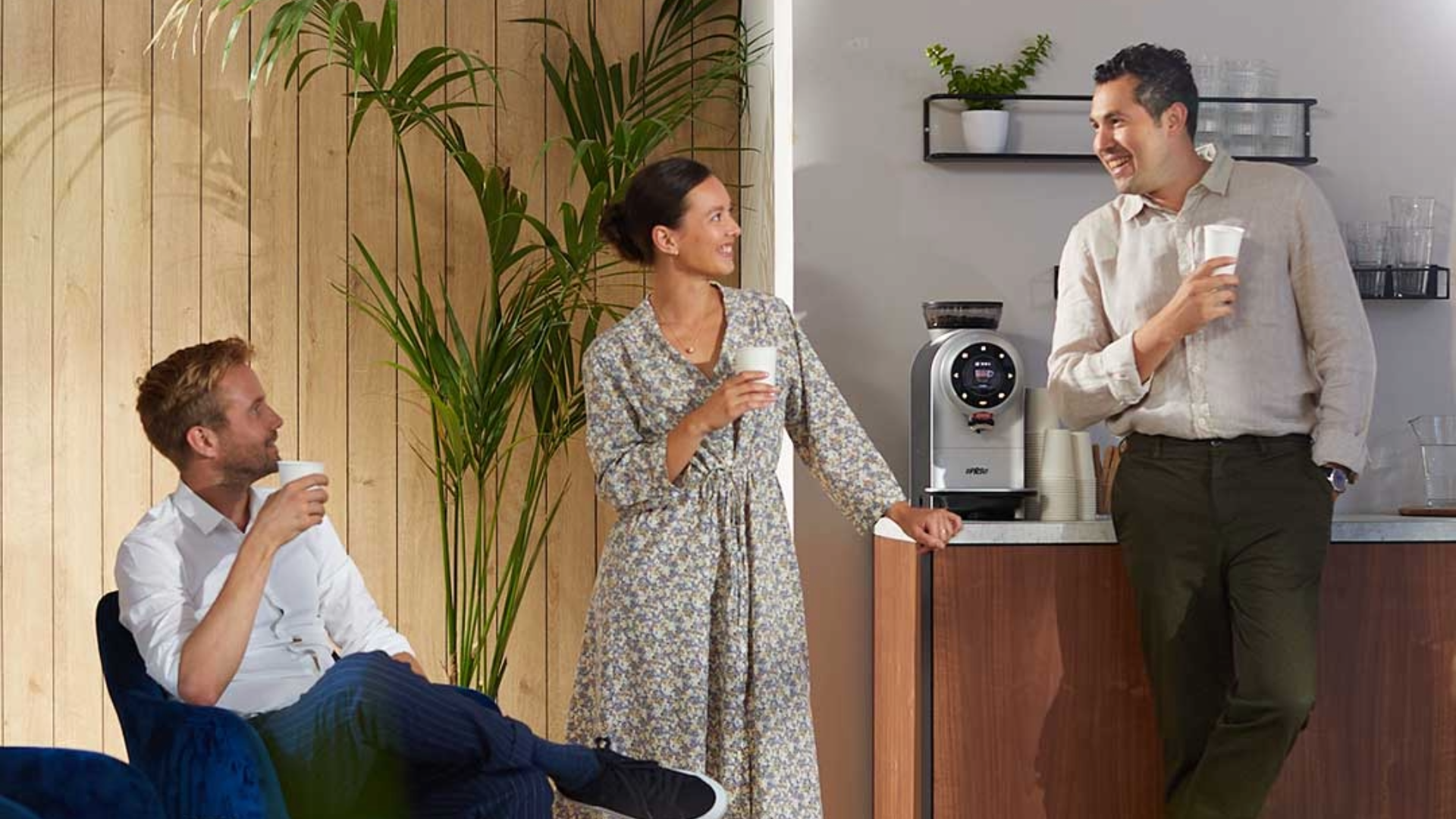 Drei Personen mit Kaffee in einem Pausenraum mit Kaffeemaschine und Pflanzen.